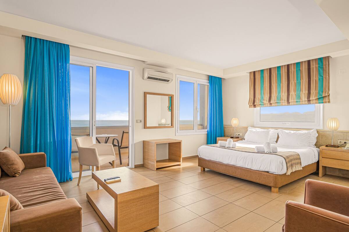 A hotel room with a bed, a sofa, a desk, and a chair. There are curtains, a window, and a mirror on the wall.