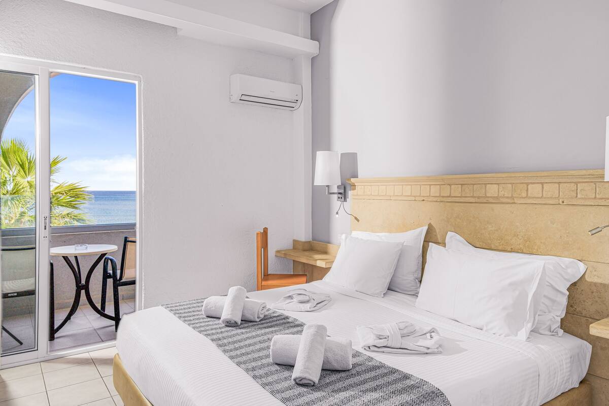 A hotel room with a bed, a small table, a chair, a balcony with a view of the sea, and an air conditioning unit.