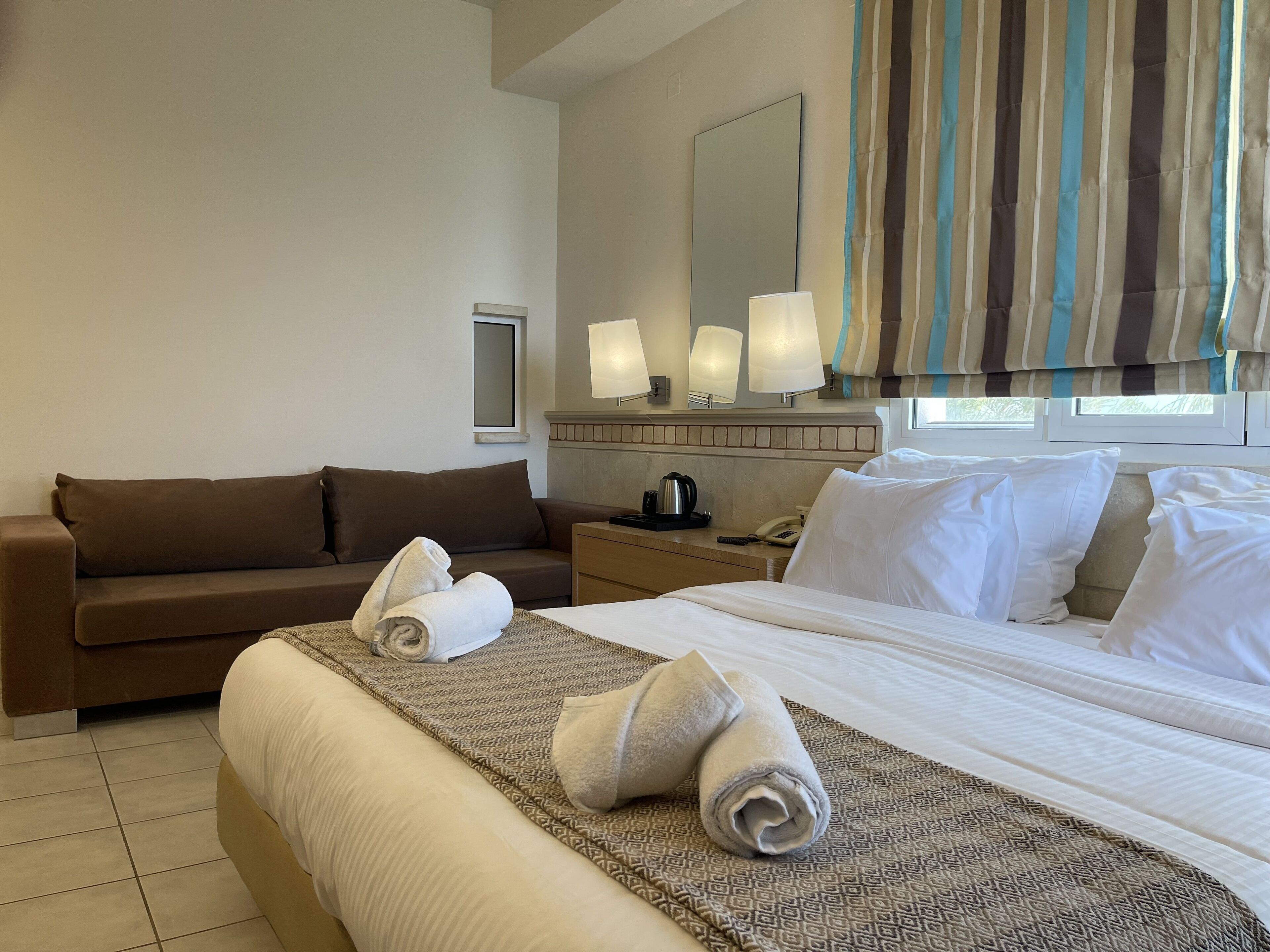 A hotel room with a bed, a sofa, a bedside table with a lamp, a mirror, and a window with curtains.