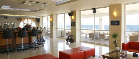 A modern hotel lobby with a bar area, red seating, and large windows offering ocean views.