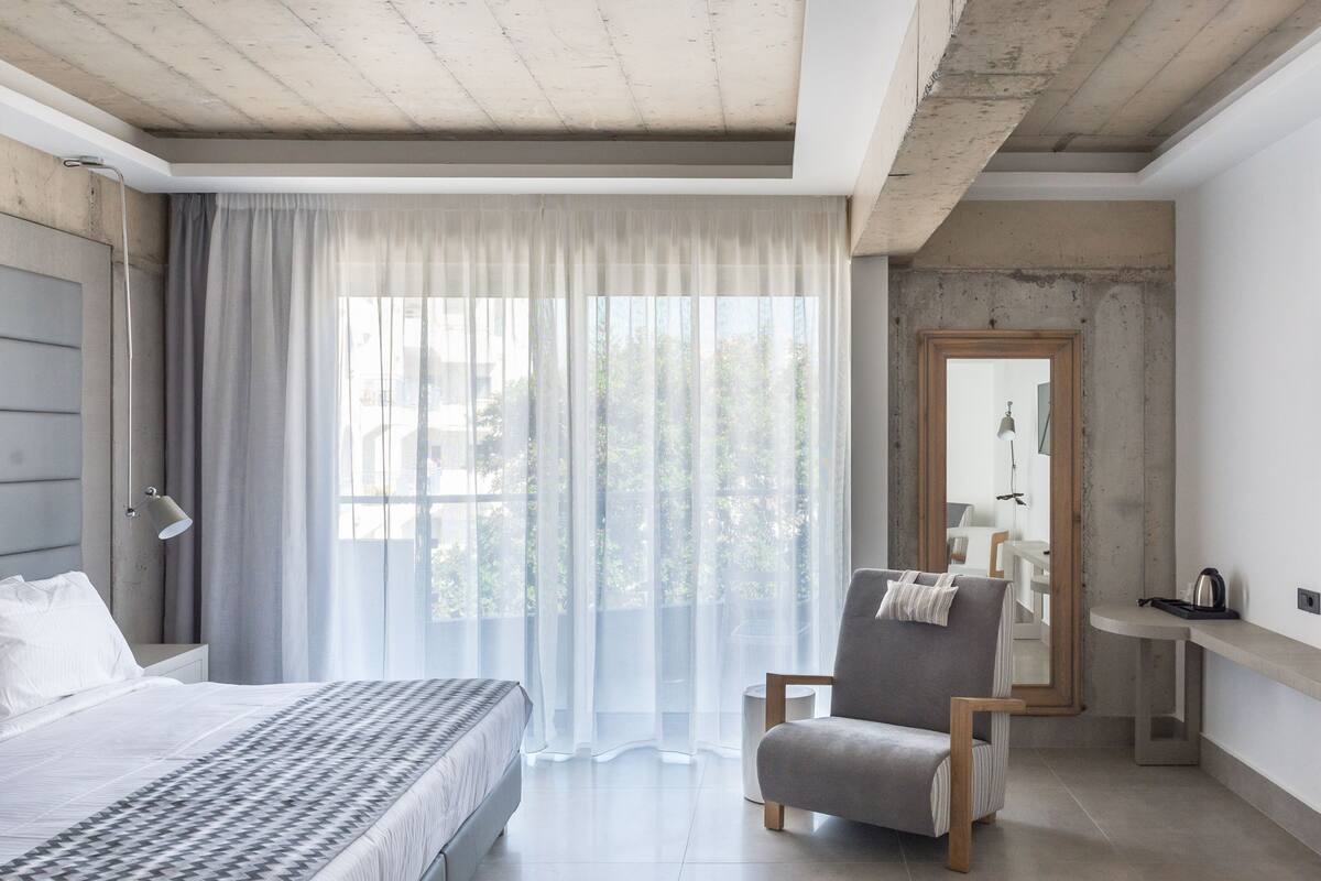 A modern hotel room with a large bed, a chair, a desk, and a view of greenery through sheer curtains.