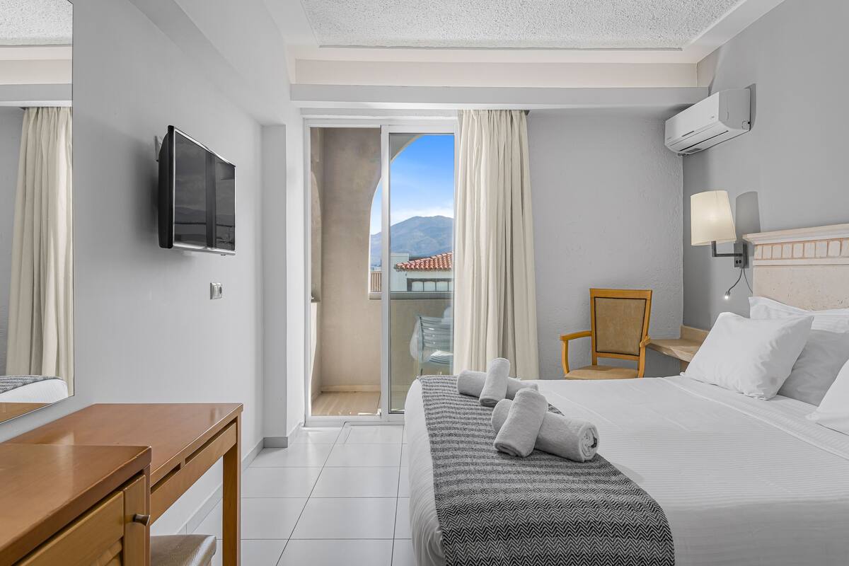 A hotel room with a bed, a desk, a chair, a TV, and a view of a balcony with a mountain in the background.