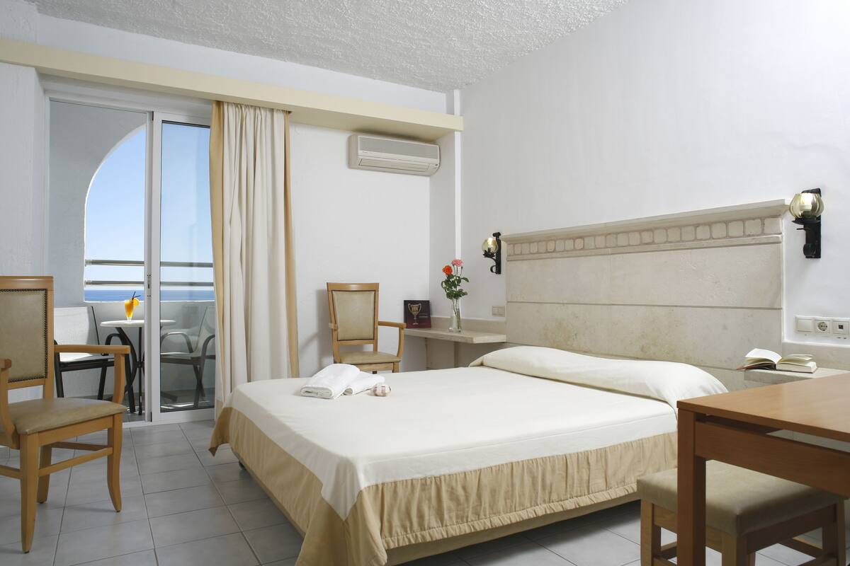 A hotel room with a bed, a desk, a chair, and a view of the sea through a sliding door.