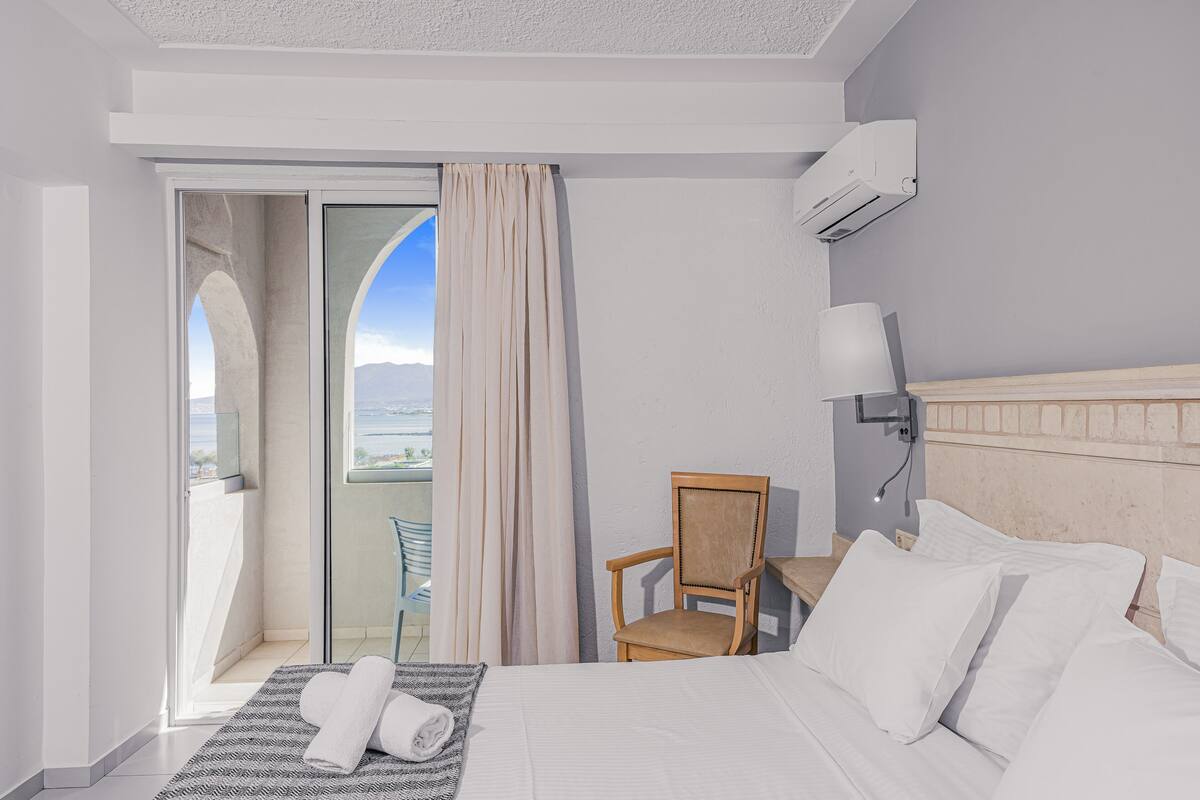 A hotel room with a bed, a chair, a table, a window with curtains, and an air conditioning unit.