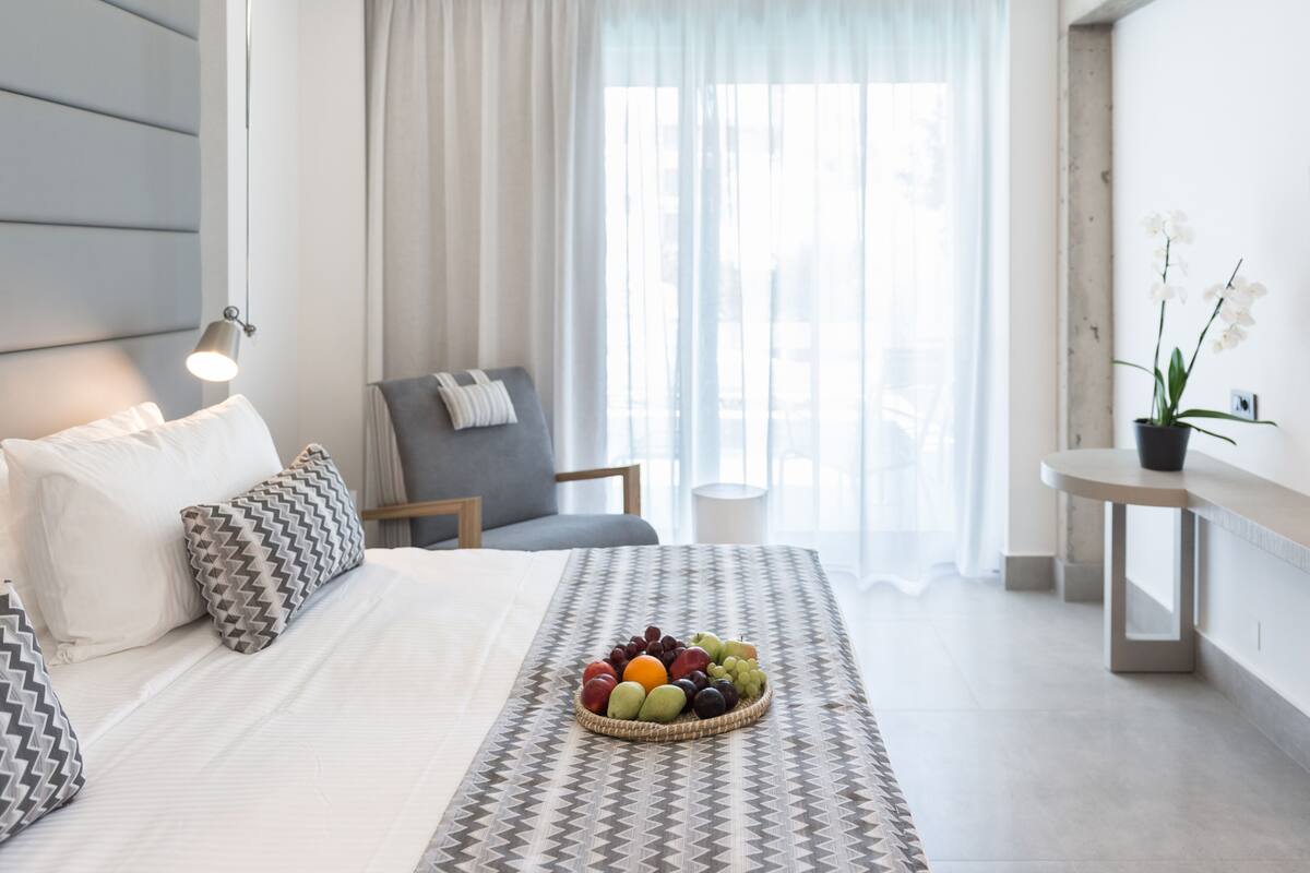 A modern hotel room with a large bed, a seating area with a chair and a small table, a fruit bowl on the bed, and a potted plant on a side table.