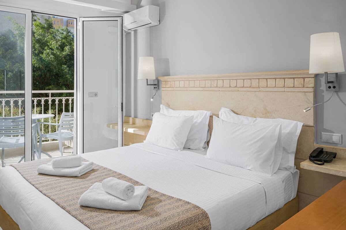 A hotel room with a bed, a bedside table with a lamp, a phone, and a view of the outside.