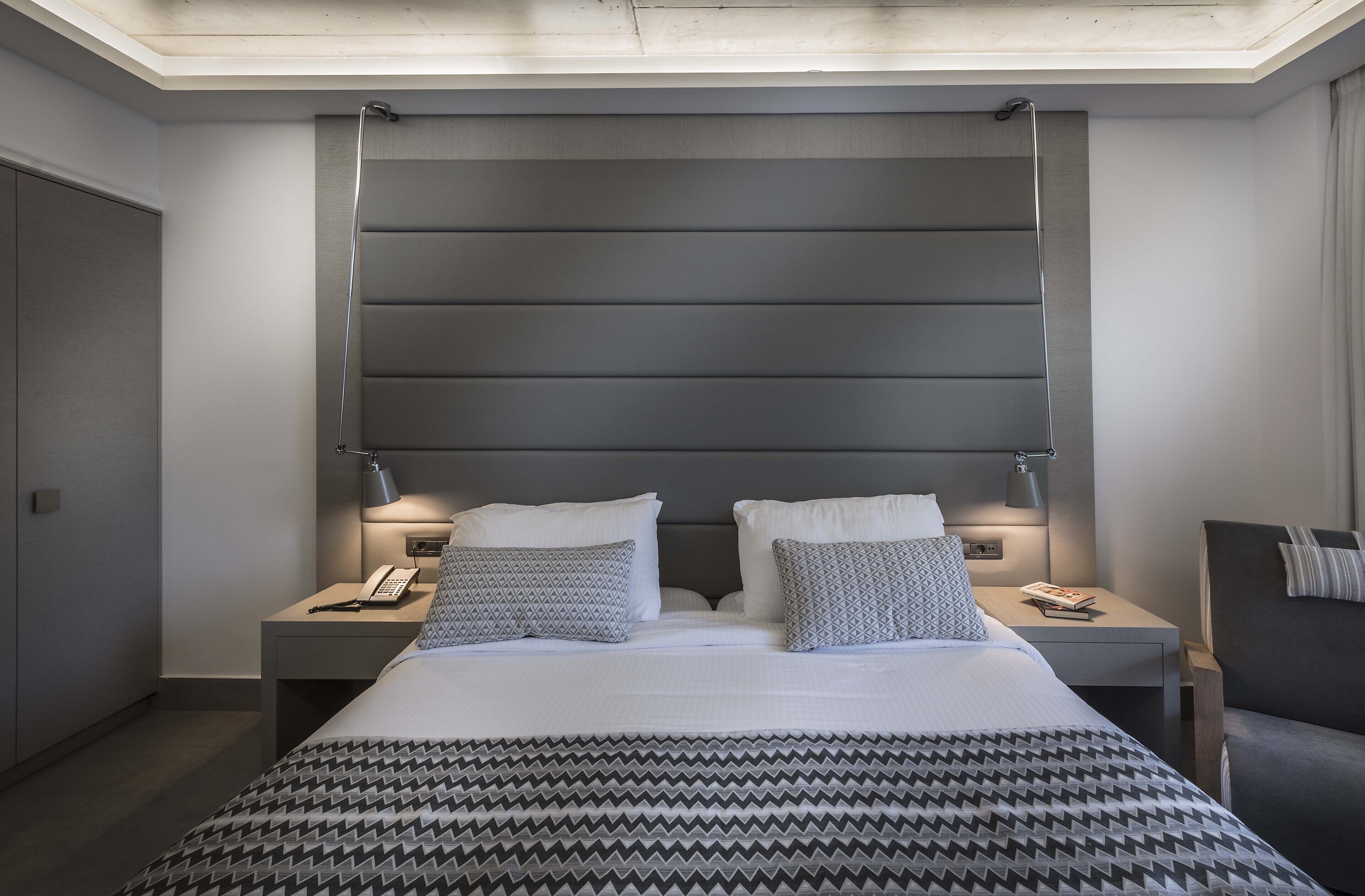 A modern hotel room with a large bed, two bedside tables, a nightstand, a chair, and a headboard with built-in lighting.