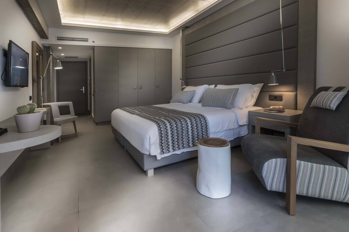 A modern bedroom with a bed, bedside tables, a chair, a small table with a plant, and a TV on the wall.