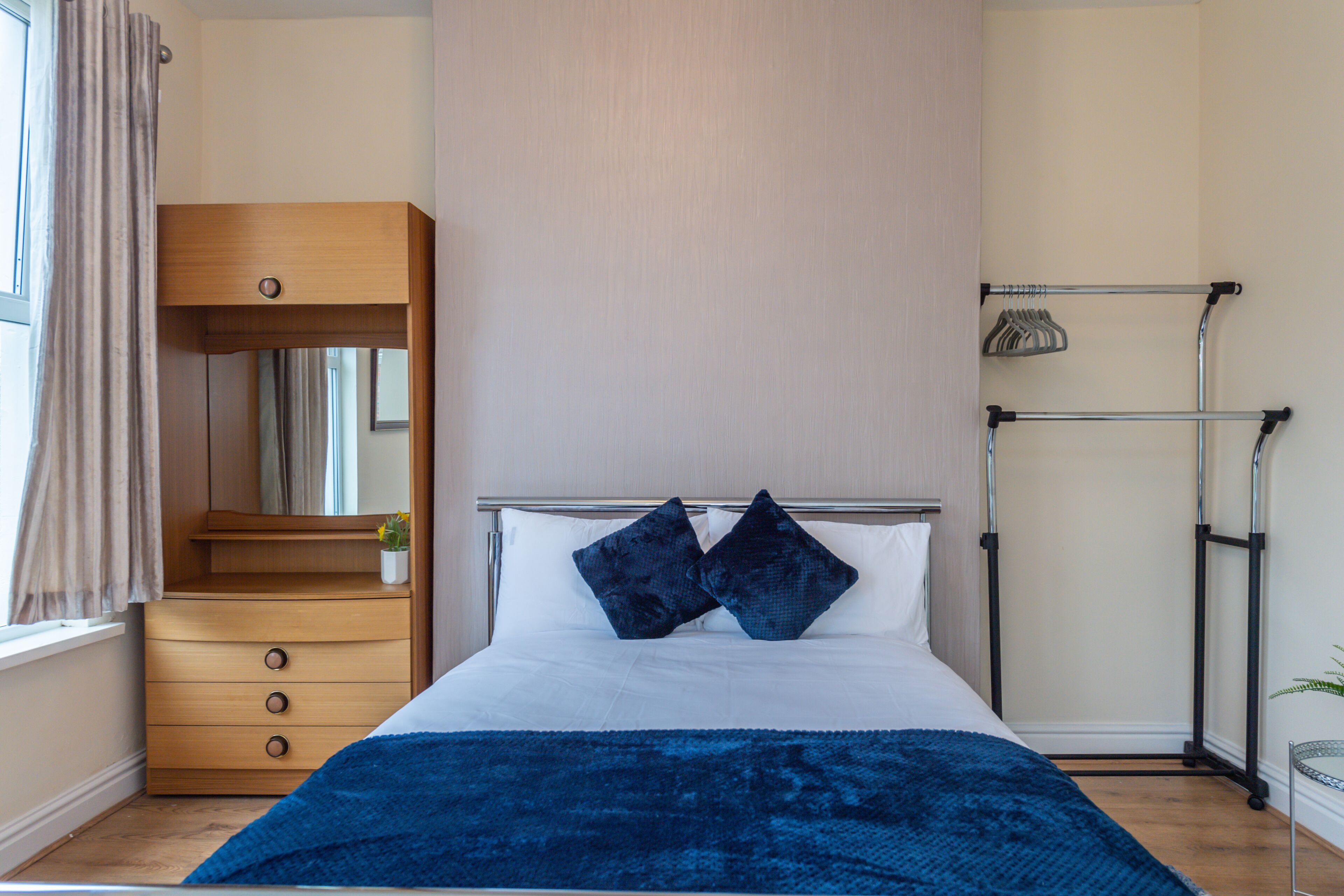A bedroom with a bed, a wooden dresser, a mirror, and a drying rack.
