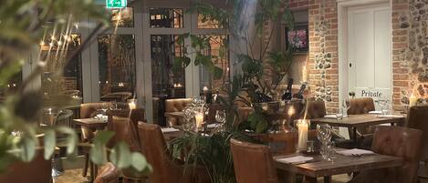 A restaurant interior with leather chairs, tables set with wine glasses, candles, and a large plant centerpiece.