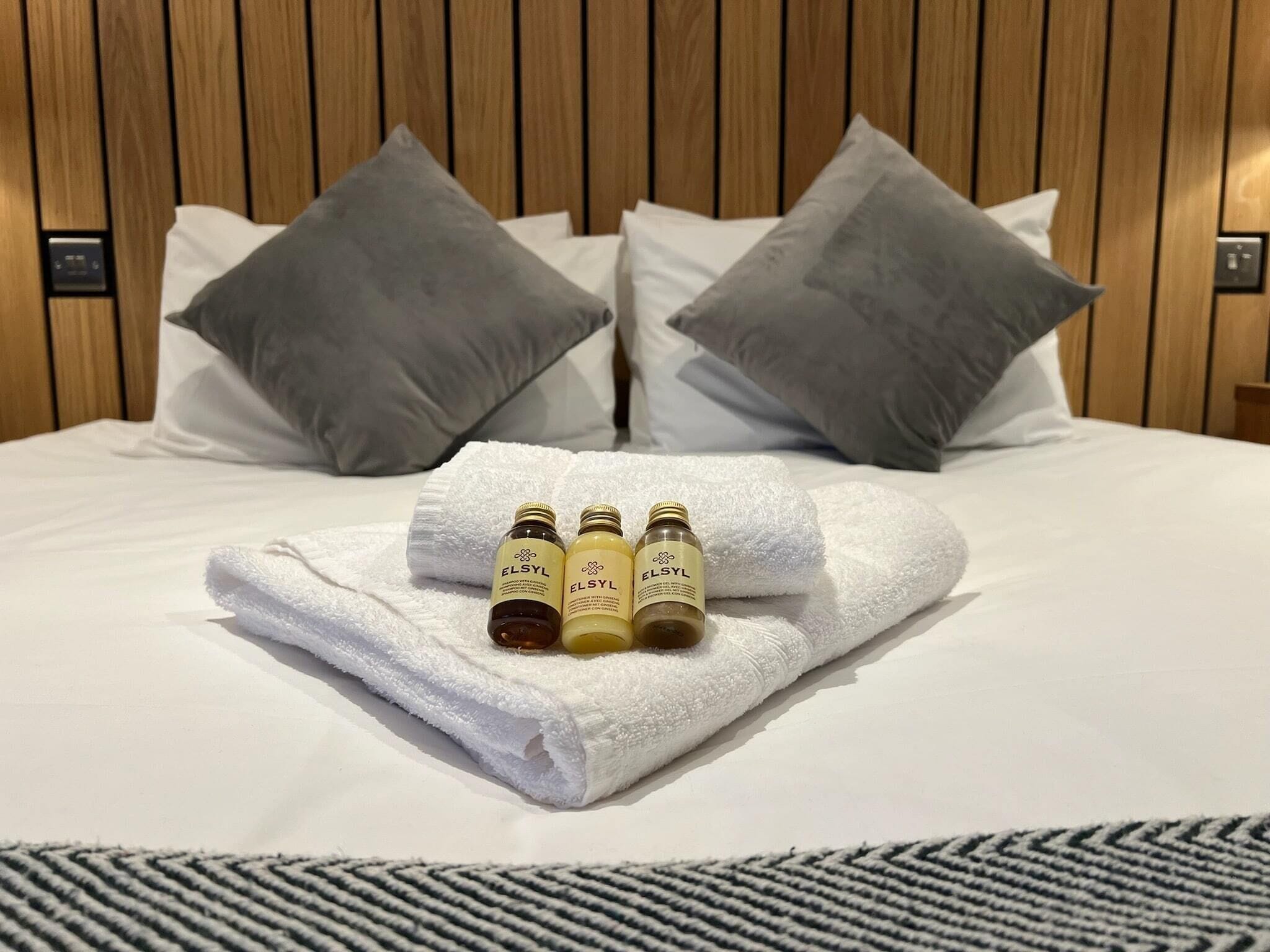 A neatly made bed with a folded white towel and two bottles of ELSYL on top.