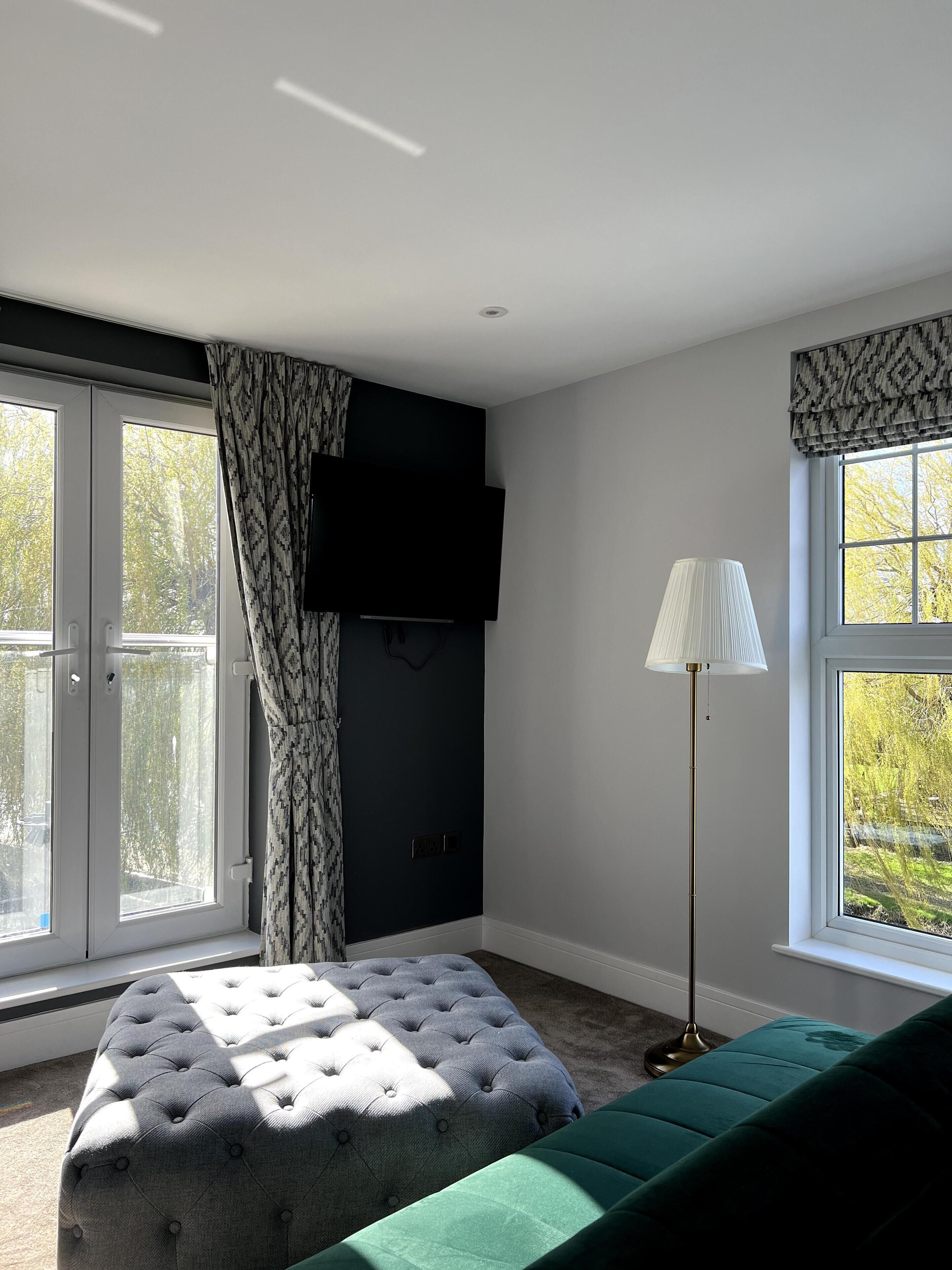 A modern bedroom with a large window, a tufted ottoman, a green sofa, a floor lamp, and a mounted TV.