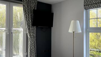 A modern bedroom with a large window, a tufted ottoman, a green sofa, a floor lamp, and a mounted TV.