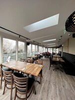 Uma sala de jantar espaçosa com pé-direito alto, grandes janelas e diversas opções de assento, incluindo mesas e bancos.