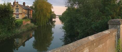 Un río tranquilo con un puente de piedra, un sauce llorón y un edificio con un letrero que dice "The Three Pickles".