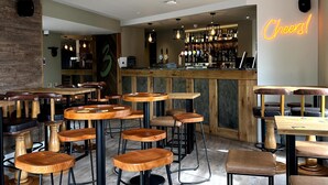 Angolo bar moderno con tavoli e sgabelli in legno, un cartello luminoso con scritto 'Cheers!' e un bancone fornito di bottiglie.