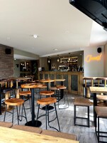 Uma área de bar moderna com mesas e banquetas de madeira, um letreiro de neon com a palavra 'Cheers!' e um balcão repleto de garrafas.