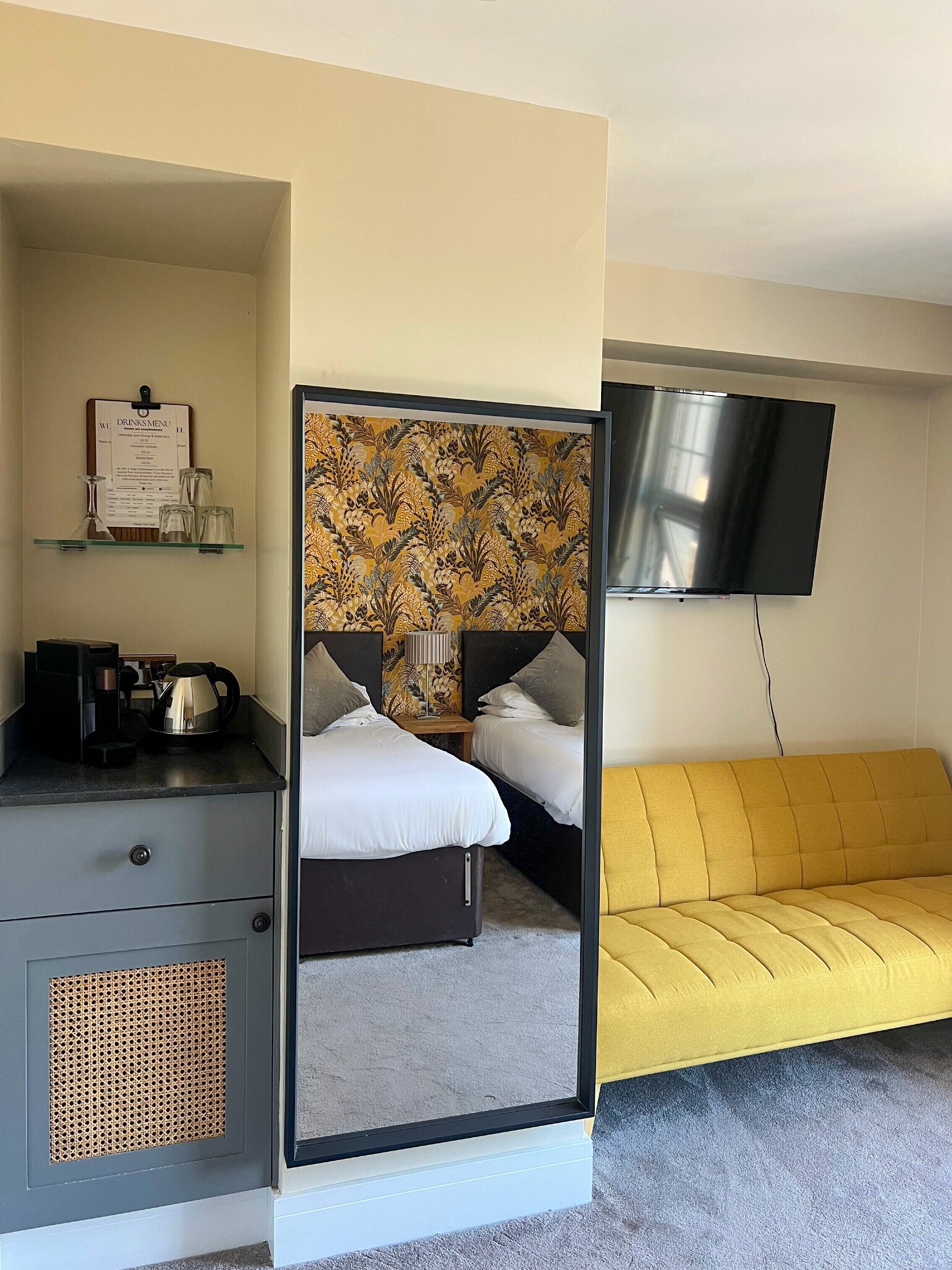 Una habitación de hotel con cama, mesita de noche con cafetera, televisor montado en la pared y sofá amarillo.