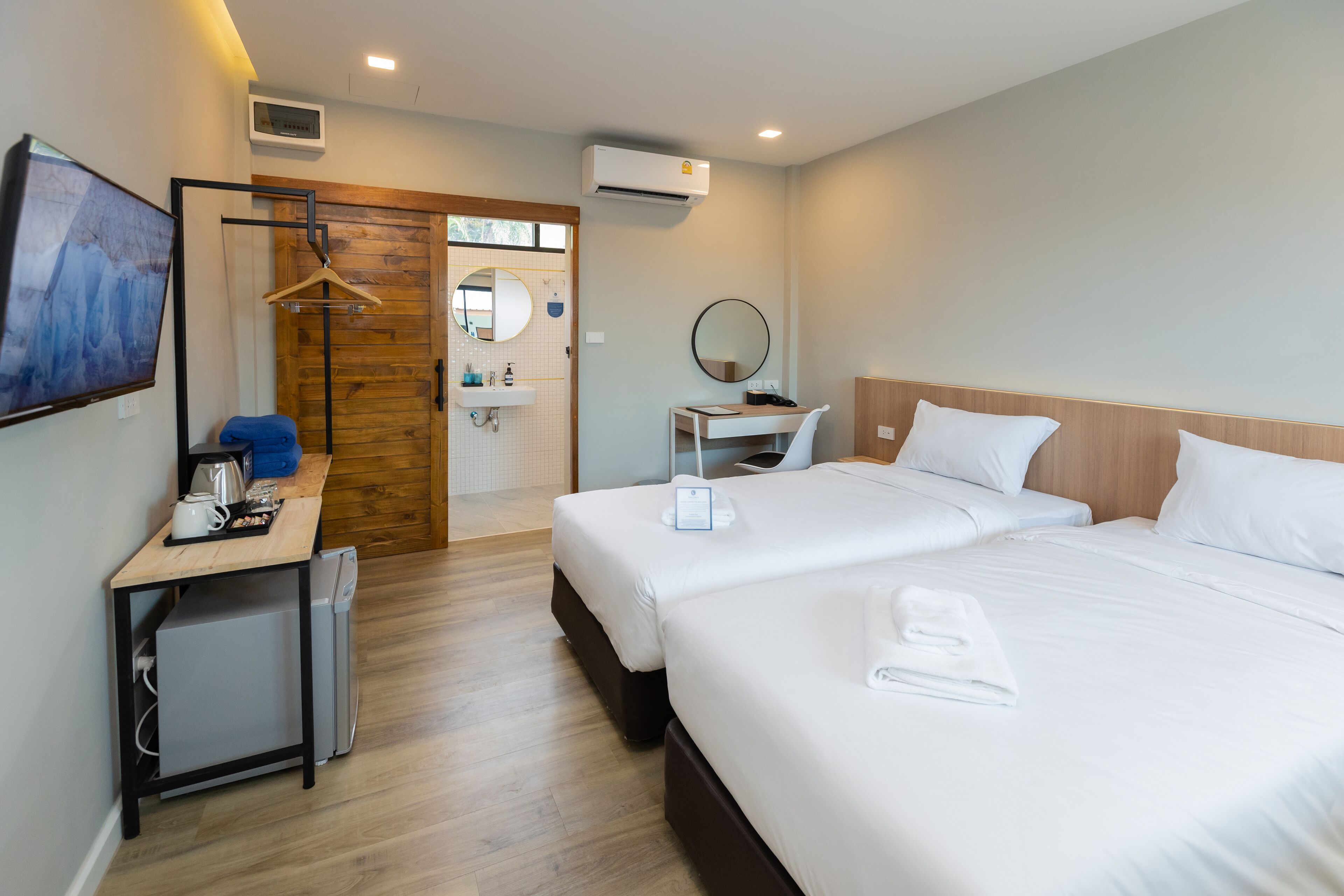 Una habitación de hotel con dos camas, un escritorio con computadora, un refrigerador pequeño, una puerta de madera y un baño con lavamanos y espejo.
