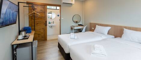 Una habitación de hotel con dos camas, un escritorio con computadora, un refrigerador pequeño, una puerta de madera y un baño con lavamanos y espejo.