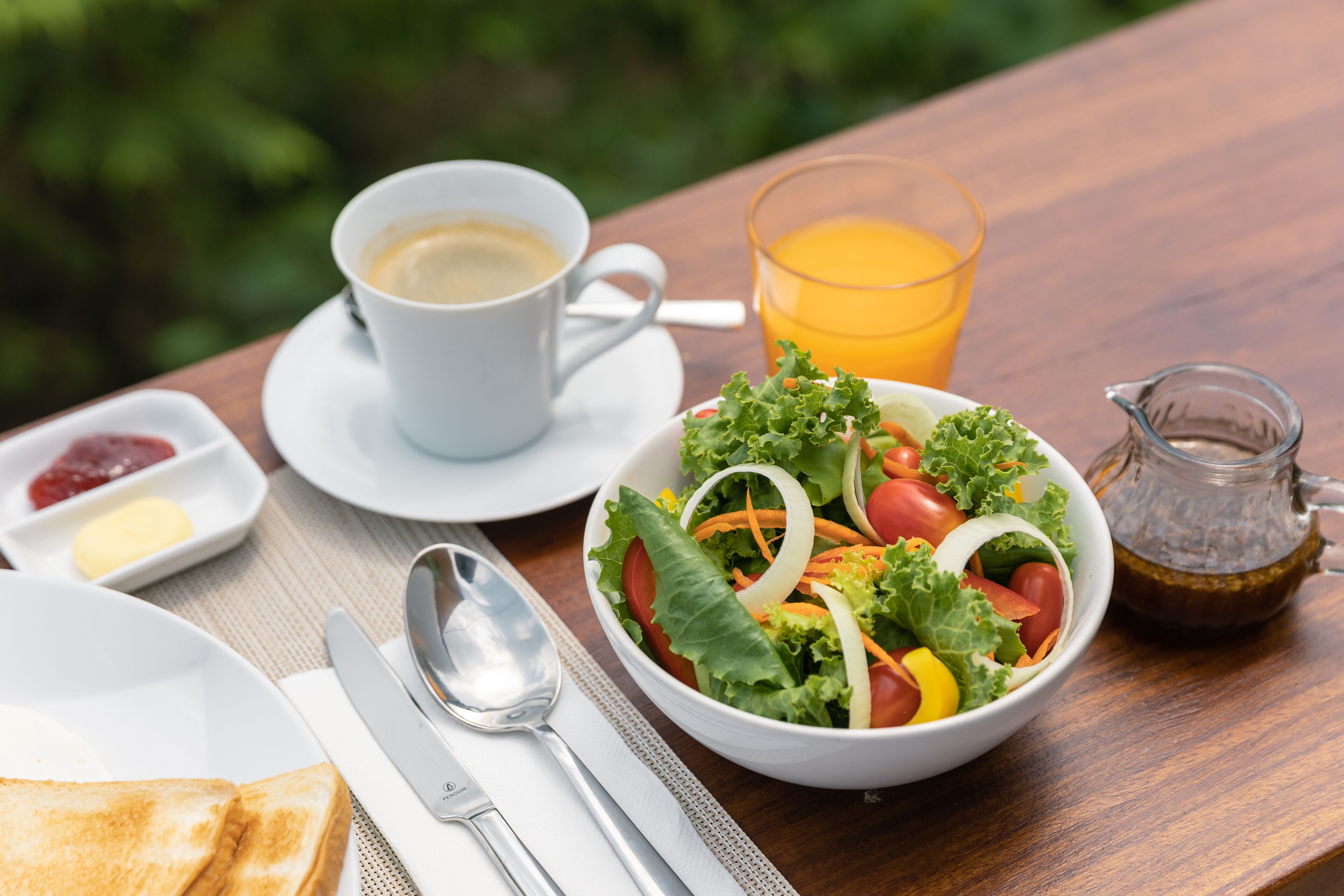 A breakfast setting with a bowl of salad, a cup of coffee, a glass of orange juice, toast, butter, jam, and a pitcher of tea on a wooden table.