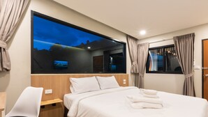 Habitación de hotel con un ventanal grande, cama con ropa blanca, escritorio con silla y televisor en la pared.
