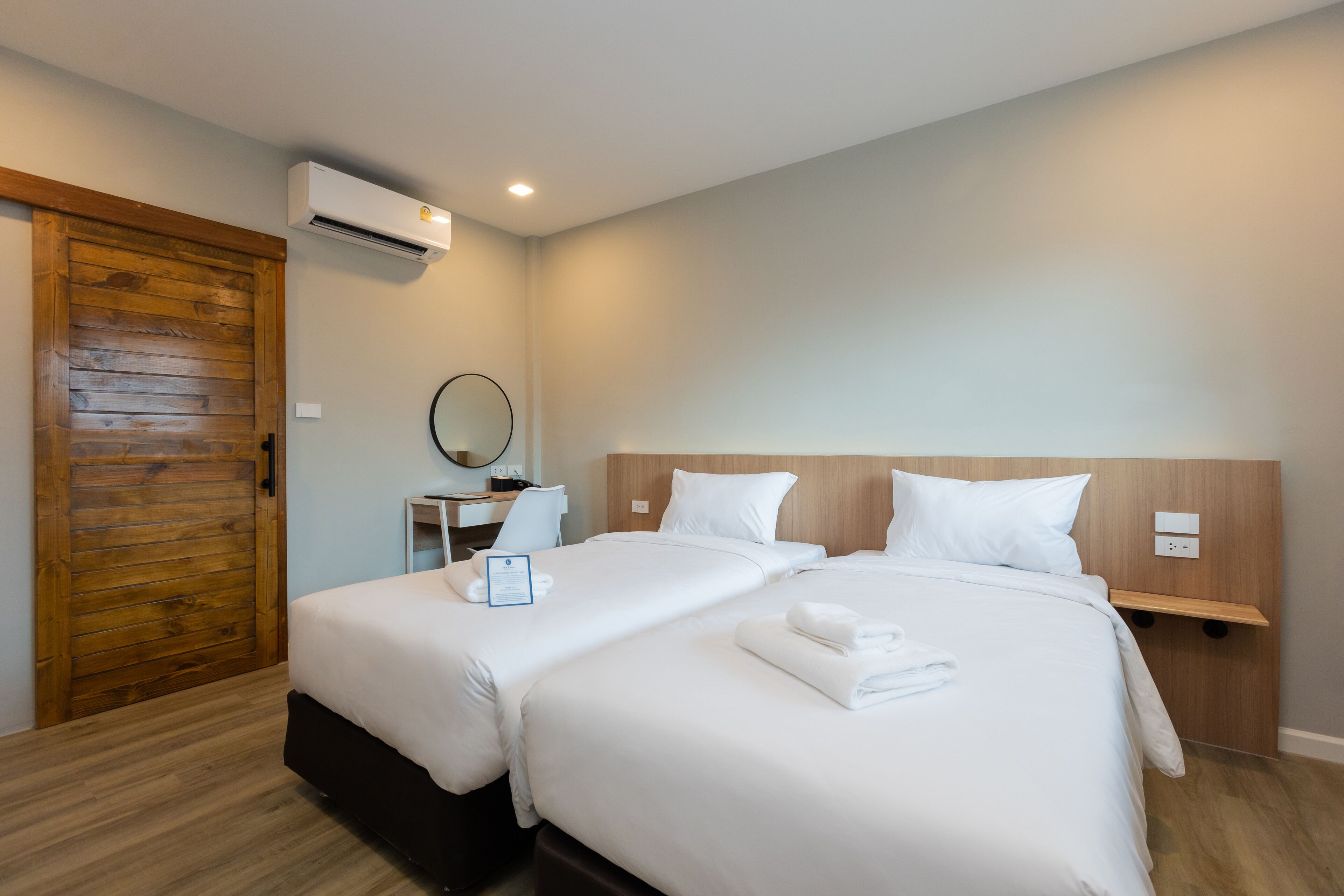A hotel room with two beds, a wooden door, a desk with a chair, a round mirror, and an air conditioning unit.