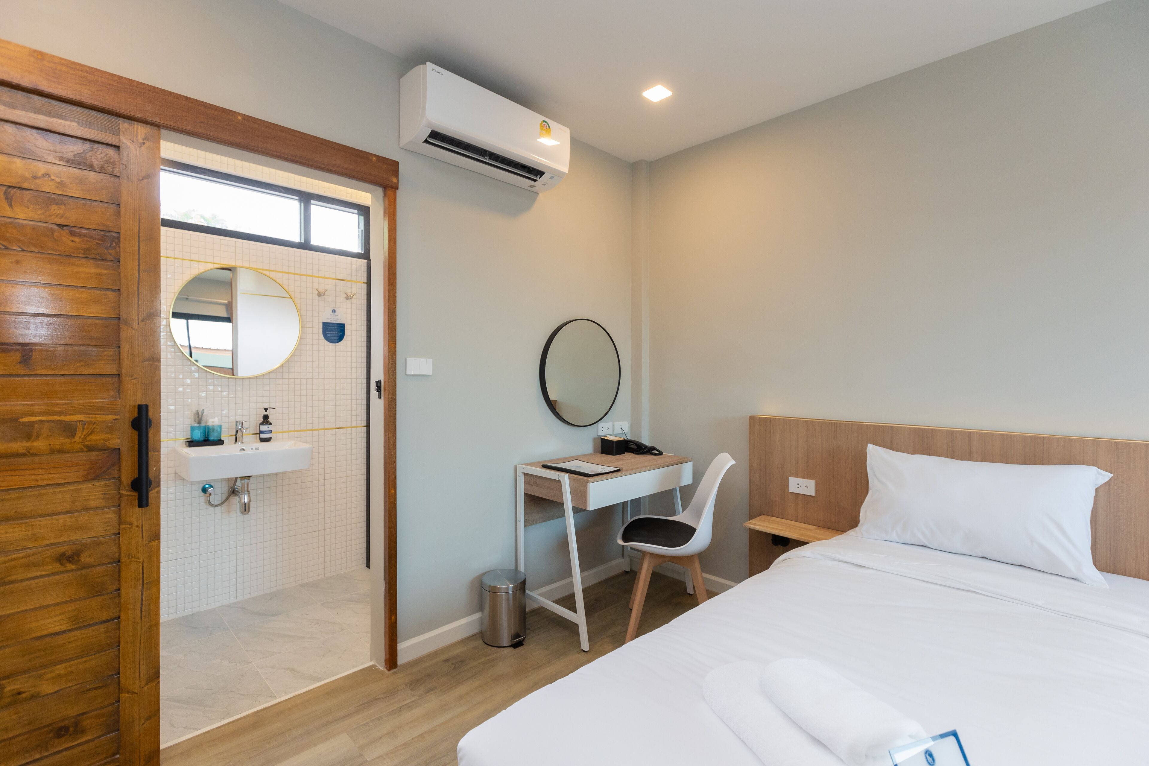 A hotel room with a wooden door, a desk with a chair, a bed, a round mirror, and a small trash bin.