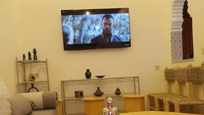 En hyggelig stue med et TV ophængt på væggen, en sofa, et sofabord med glas og en tepotte, samt en dekorativ vægdekoration.