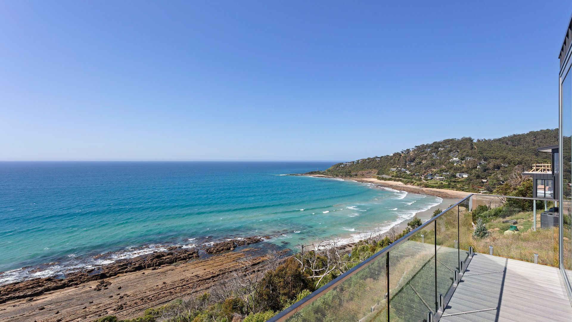 A coastal view from a balcony overlooking a beach with clear blue waters and a hilly landscape in the background.