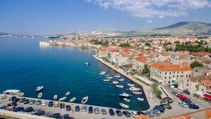 Une ville côtière dotée d’un port de plaisance rempli de bateaux, d’une rangée de bâtiments aux toits recouverts de tuiles rouges et offrant une vue dégagée sur la mer et les montagnes lointaines.