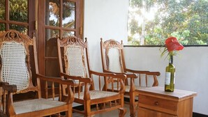A wooden chair with a woven seat, a wooden table with a bottle and a red flower, and a window with a view of greenery. - Charming 2-bed Apartment in Weligama (Weligama)