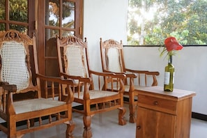A wooden chair with a woven seat, a wooden table with a bottle and a red flower, and a window with a view of greenery. - Charming 2-bed Apartment in Weligama (Weligama)