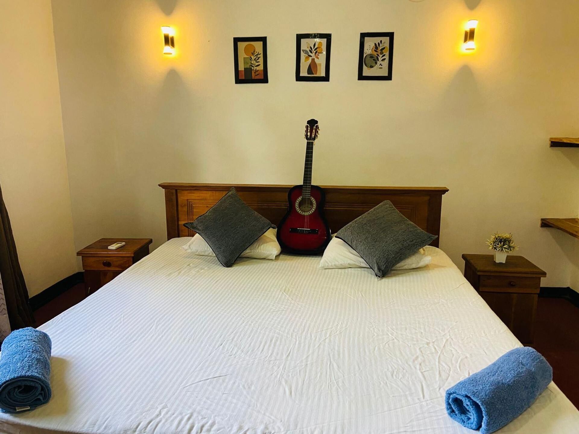 A hotel room with a large bed, a guitar, two pillows, a bedside table, a nightstand, and wall-mounted lights.