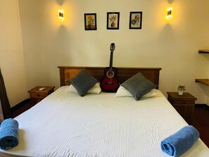 Habitación de hotel con una cama grande, una guitarra, dos almohadas, una mesita de noche y luces fijadas en la pared.