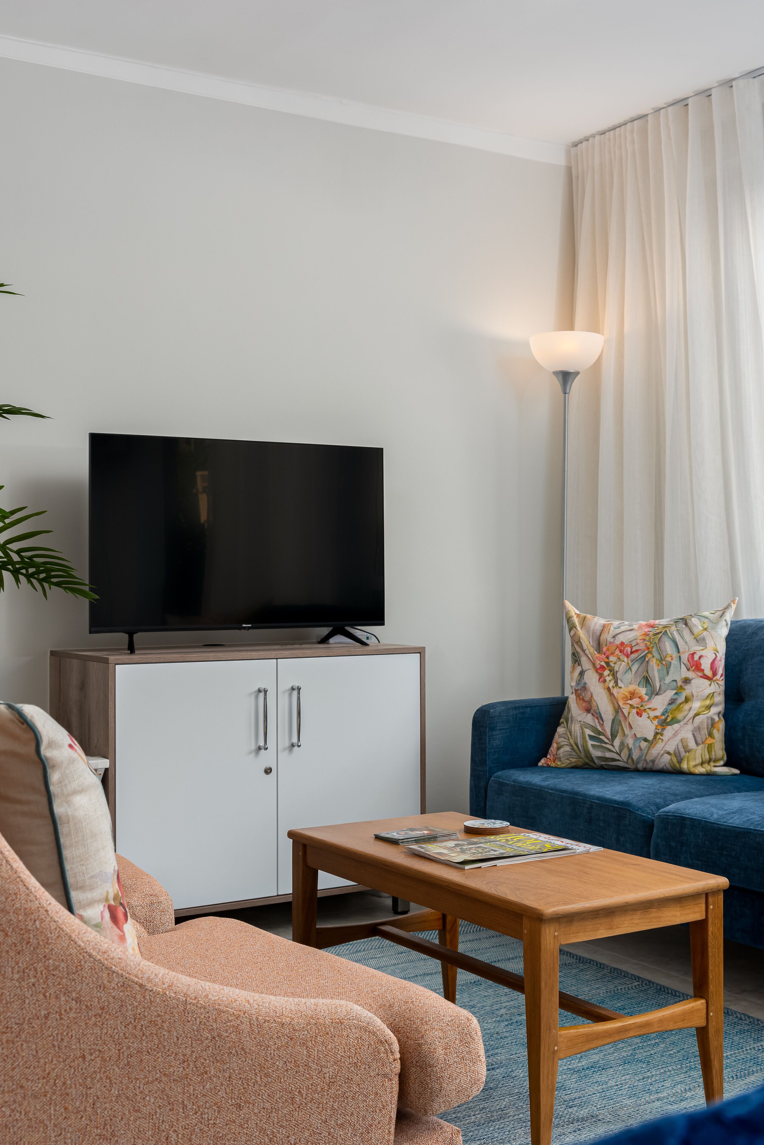 Een woonkamer met een televisie op een kast, een blauwe bank, een houten salontafel en een vloerlamp.