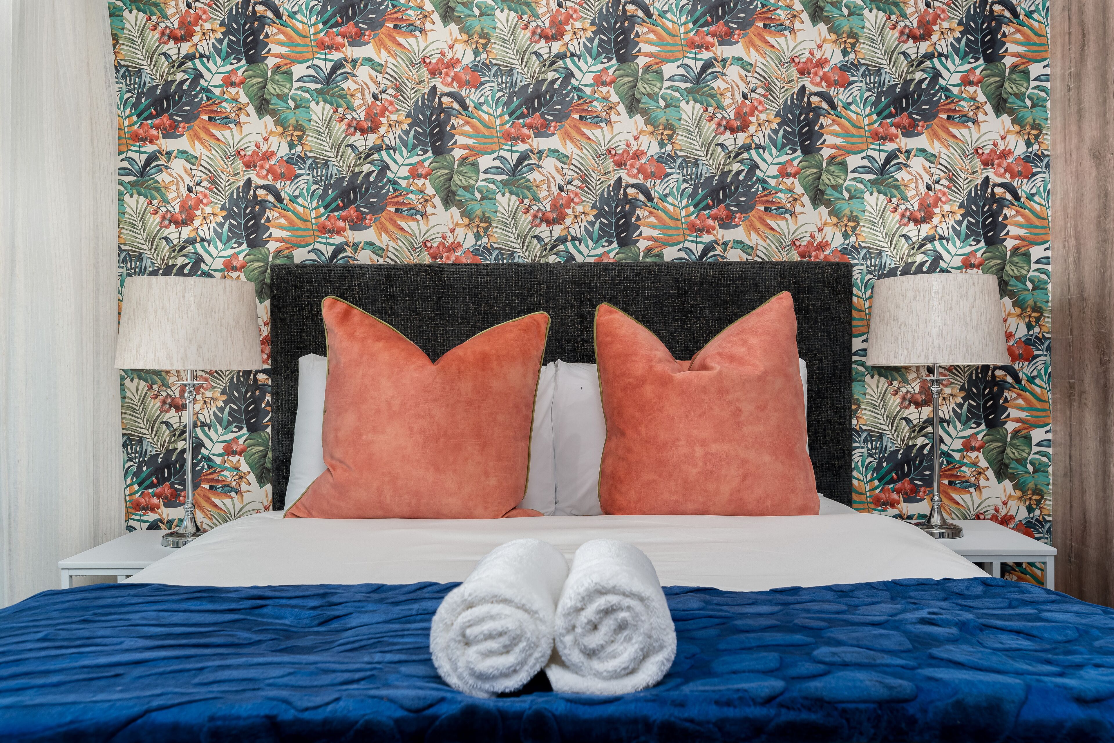 A bed with a blue bedspread, two orange pillows, and two white towels.