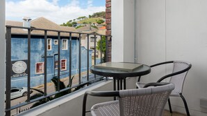 Un balcon avec une table en verre et des chaises en osier, donnant sur une rue bordée d’immeubles et sur une colline en arrière-plan.