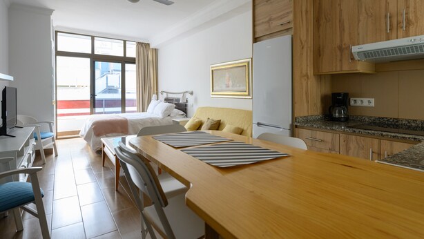 Une chambre d’hôtel moderne avec une table à manger en bois, un lit avec une tête de lit beige, une kitchenette équipée d’un réfrigérateur et d’une machine à café, et une grande fenêtre avec des rideaux.