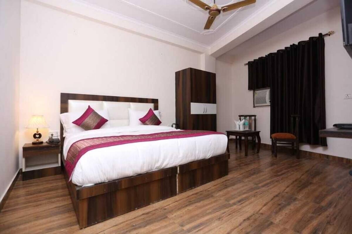 A hotel room with a large bed, a nightstand with a lamp, a wooden floor, a ceiling fan, a small table with a chair, and a window with curtains.