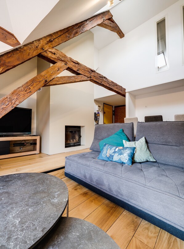 Una sala de estar moderna con vigas de madera a la vista, un sofá gris con cojines estampados, un televisor de pantalla plana y una chimenea.