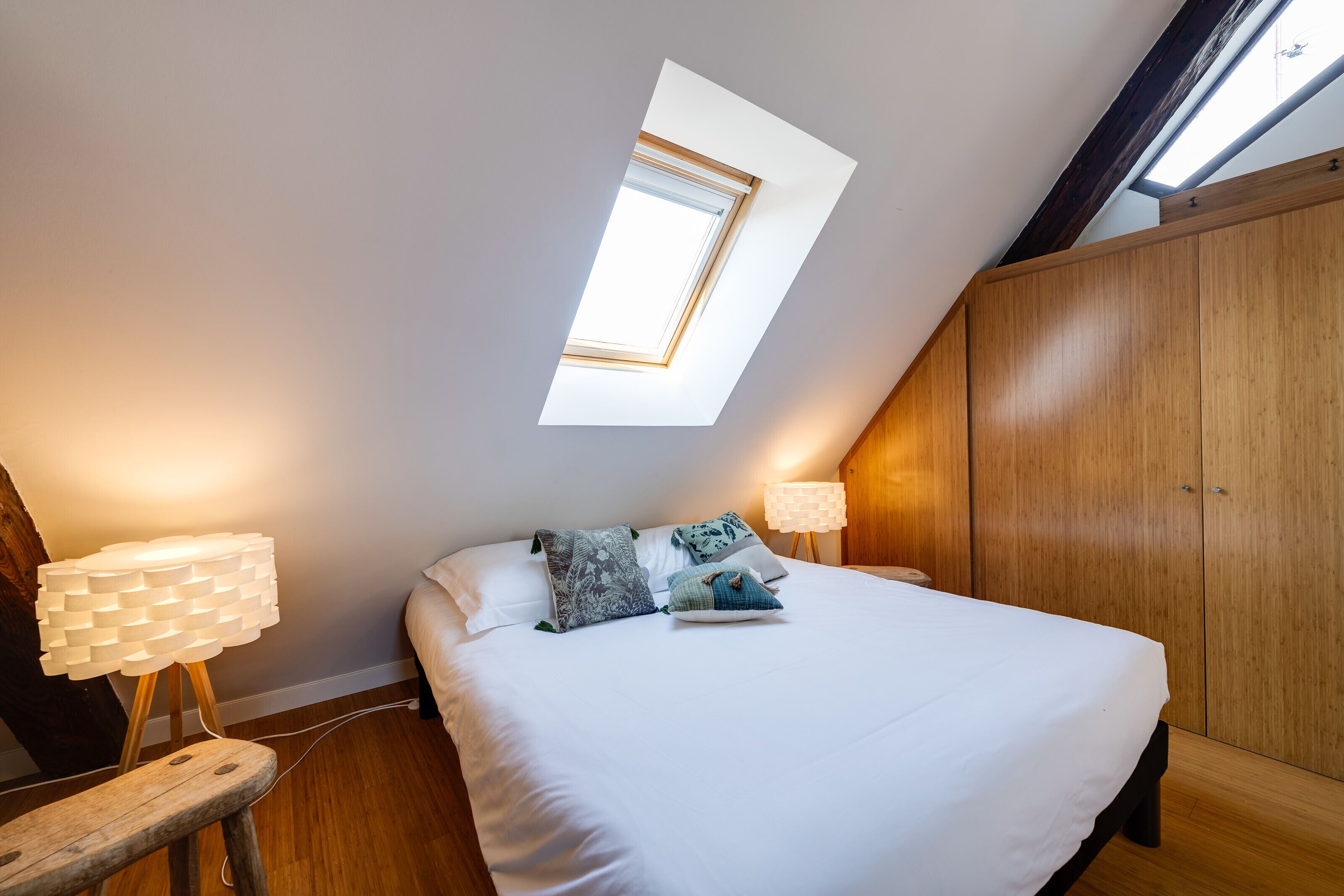 A bedroom with a skylight, a bed with white bedding, two bedside lamps, and a wooden wardrobe.