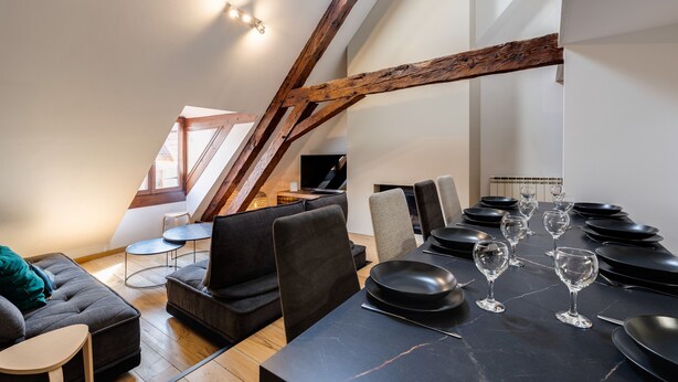 A modern loft-style room with a dining area, high ceilings, exposed wooden beams, and a cozy seating arrangement.