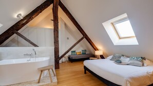 A bedroom with a large bed, a nightstand, a bedside table, a window, a skylight, and a bathroom with a bathtub.