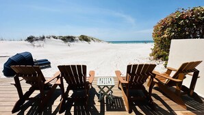 A beachside deck with wooden chairs and a table, overlooking a sandy beach and ocean.