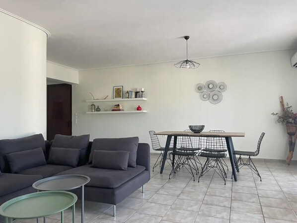 Un salon moderne avec un canapé gris, une petite table ronde et un coin repas comprenant une table en bois et des chaises en fil de métal.
