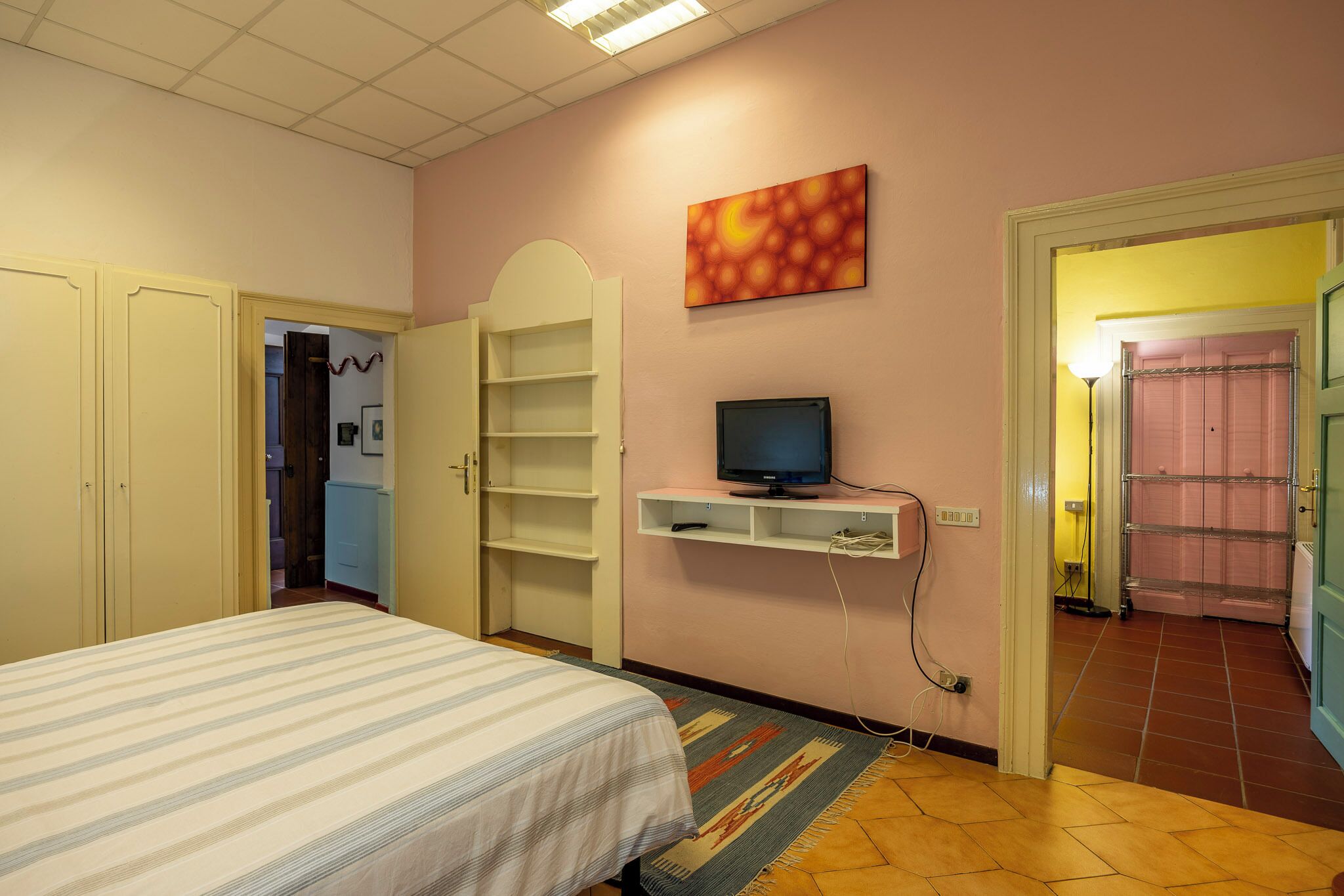 Une chambre à coucher avec un lit, un téléviseur posé sur une étagère, une armoire et une porte de salle de bain visible au fond.