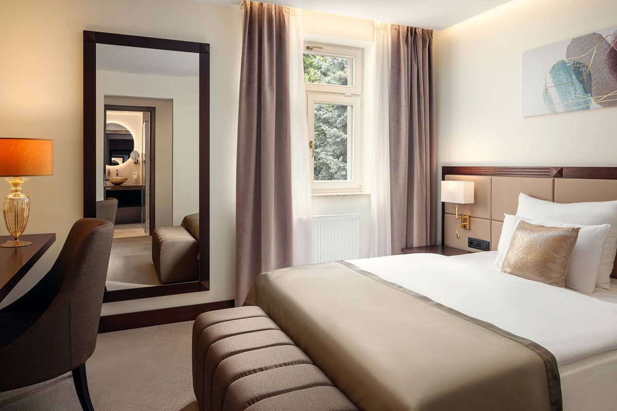 Habitación de hotel con una cama grande, un escritorio con lámpara, una silla, un espejo y una ventana con cortinas.