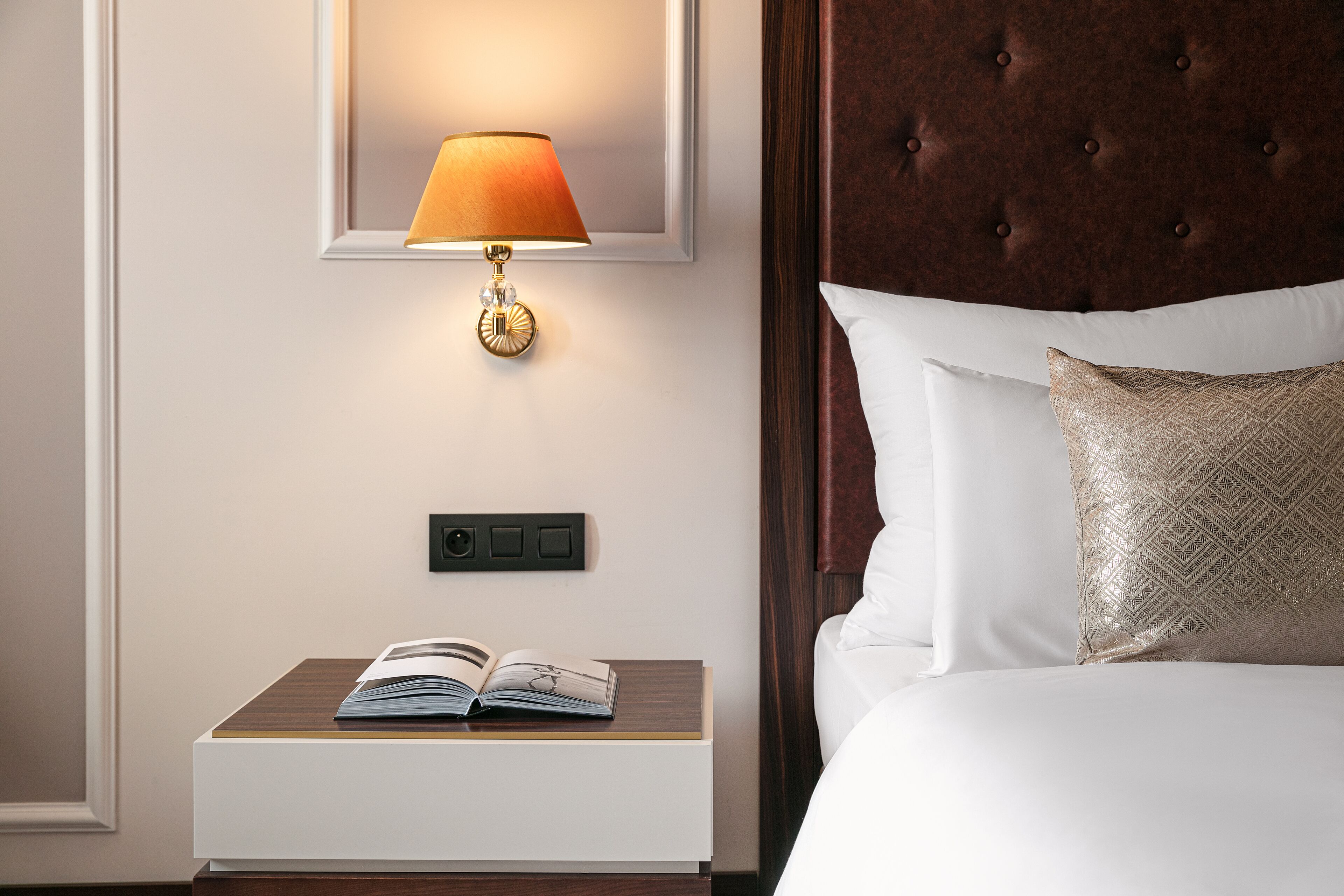 A neatly made bed with white linens, a gold-patterned pillow, and a book on a bedside table.