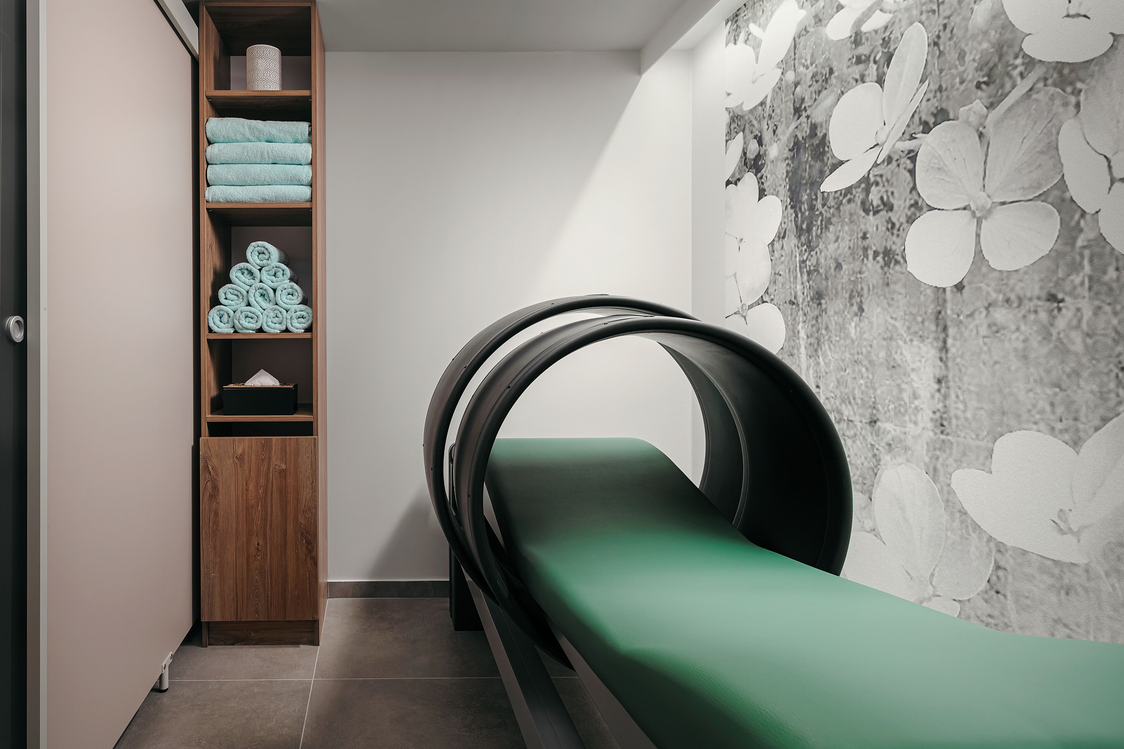 A room with a green bed, a wooden shelf with towels, and a wall with a floral pattern.